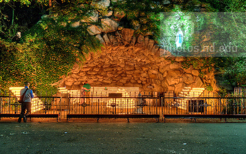 the Grotto at night with many candles lit. A student kneels and prays at a railing in front of the Grotto